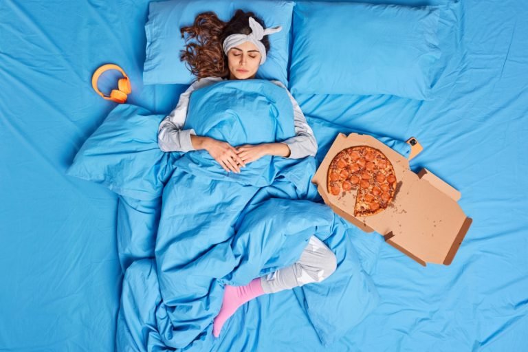 Calm millennial woman takes nap lies under soft blanket eats delicious pizza stays in bed enjoys goo